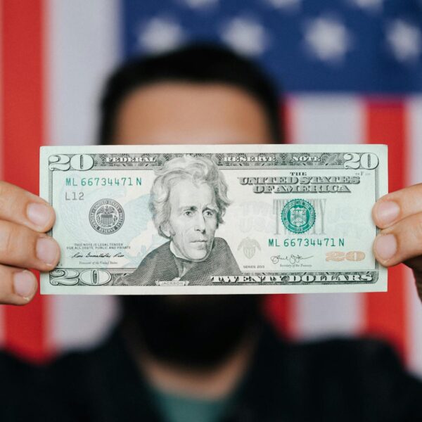 Close-up of a man holding a 20-dollar bill with an American flag blurred in the background, symbolizing finance and patriotism.