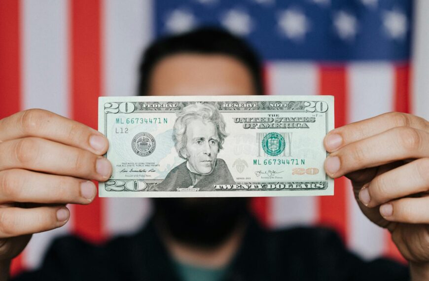 Close-up of a man holding a 20-dollar bill with an American flag blurred in the background, symbolizing finance and patriotism.